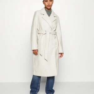 Beige kappa med bälte - Snygg beige kappa med knytbälte i midjan och klassisk krage. Perfekt för att hålla stilen under kyligare dagar. Långärmad design som ger en elegant look. Använt 3 gånger, köpt för 700kr säljer den för 500. Kan mötas upp!