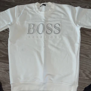 Vit sweatshirt från Hugo Boss - Säljer en stilren vit sweatshirt från Hugo Boss med ett stort BOSS-tryck på framsidan. Tröjan har långa ärmar och en rund halsringning. Perfekt för en avslappnad och trendig look. 🖤