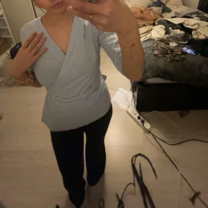 Randig omlottopp - Säljer en stilren randig omlottopp i ljusblått och vitt. Toppen har trekvartsärmar och ett knytband i midjan för en smickrande passform. Perfekt för en chic och avslappnad look.