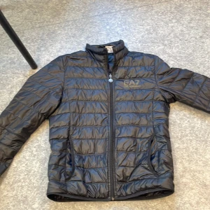 Svart pufferjacka från Emporio Armani - Säljer en svart pufferjacka från Emporio Armani med dragkedja och EA7-logga på bröstet. Jackan har en quiltad design och är perfekt för kyligare väder. Den är långärmad och har en stilren look.