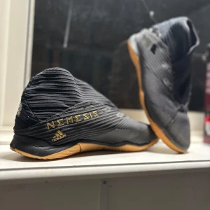 Adidas Nemesis fotbollsskor - Snygga Adidas Nemesis fotbollsskor i svart med guldiga detaljer. Skorna har en unik design med texten 'Nemesis' på sidan och en slitstark sula i gummi. Perfekta för inomhusfotboll och ger bra grepp på planen.
