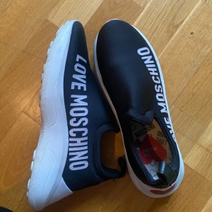 Svarta sneakers från Love Moschino - Snygga svarta sneakers från Love Moschino med vit sula och textdetaljer. Skorna har ett hjärtmönster på sulan och en röd hjärtdetalj. Stora i storlek! 