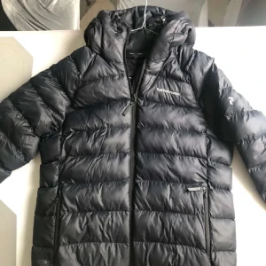 Svart dunjacka från Peak Performance - Säljer en svart dunjacka från Peak Performance. Jackan har en dragkedja framtill och är quiltad för extra värme. Den har en huva och långa ärmar, perfekt för kyliga dagar. En stilren och funktionell jacka för vintern.