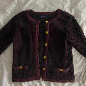 Vinröd kofta från Stockh LM - Säljer en snygg vinröd kofta från Stockh LM med guldfärgade knappar. Koftan har en rund halsringning och två dekorativa fickor framtill. Perfekt för att ge en elegant touch till din outfit.