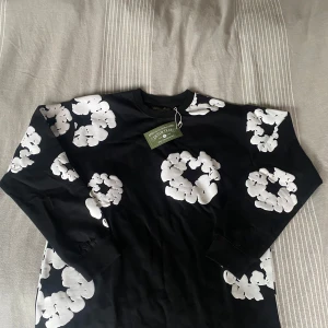 Svart tröja från Denim Tears - Säljer en svart tröja från Denim Tears med ett iögonfallande vitt blommönster. Tröjan är tillverkad i 100% bomull och har långa ärmar. Perfekt för dig som vill ha något unikt i garderoben!