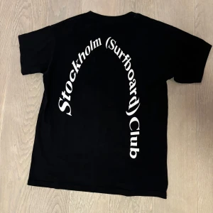 Stockholm Surfboard Club T-shirt - Snygg svart t-shirt med texten 'Stockholm (surfboard) Club' i vitt tryck på baksidan. Perfekt för en avslappnad stil med en touch av surfkultur.