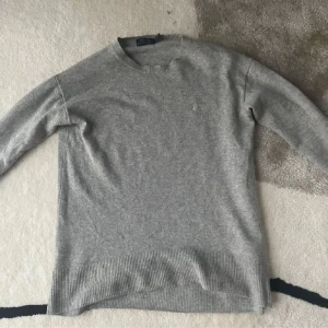 Grå merinoulltröja från Ralph Lauren - Säljer en stilren grå tröja från Ralph Lauren i mjuk merinoull. Tröjan har en diskret broderad logga på bröstet och ribbade kanter vid ärmslut och nederkant. Skick 9/10! Storlek XS! Skriv om du har några frågor eller funderingar!