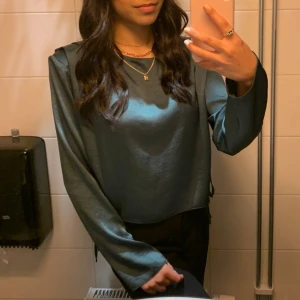 Grön långärmad blus - Snygg grön blus med långa ärmar och en elegant drapering på sidan. Perfekt för en stilren look. Materialet ser ut att vara mjukt och glansigt, vilket ger en lyxig känsla. Används bara en gång.