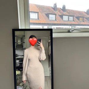 Beige ribbad stickad klänning - Säljer en stilren beige ribbad klänning med lång ärm och hög krage. Klänningen har en tight passform och en slits framtill som ger en elegant touch. Perfekt för kyligare dagar.