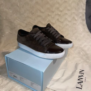  sneakers från Lanvin - Snygga bruna sneakers från Lanvin med en stilren design. Skorna har en kombination av mocka och skinn, vilket ger en lyxig känsla. De har svarta snören och en vit sula som kontrasterar snyggt mot det mörka materialet. Finns andra skosnören till. Storlek 42 men passar 43