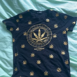 Svart t-shirt med guldtryck från Belman - Snygg svart t-shirt från Belman med ett iögonfallande guldtryck av cannabisblad och texten 'Amsterdam Officine'. Perfekt för en avslappnad stil med korta ärmar och normal passform.