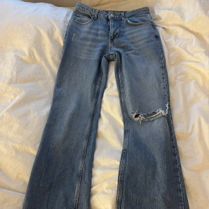 Blå jeans med slitningar - Snygga blå jeans med en cool slitning på ena benet. De har en klassisk femficksdesign och knappgylf. Perfekta för en avslappnad stil med en touch av edge.