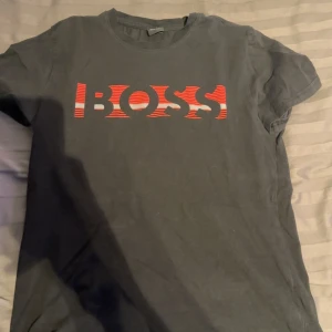 Svart t-shirt från Hugo Boss - Snygg svart t-shirt från Hugo Boss med ett coolt rött och vitt BOSS-tryck på framsidan. Perfekt för en stilren och avslappnad look. Tillverkad i mjukt material för extra komfort.