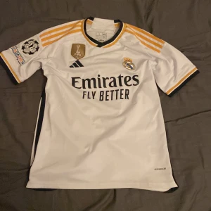 Real Madrid tröja, Mbappé 9 - Säljer en vit Real Madrid fotbollströja från Adidas med Mbappé och nummer 9 på ryggen. Tröjan har korta ärmar med guld och svarta detaljer samt klubbens emblem och sponsorlogga på framsidan. Perfekt för fans av laget och spelaren!