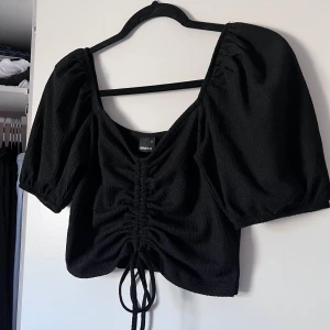Svart topp från Gina Tricot - Snygg svart topp från Gina Tricot med puffiga trekvartsärmar och en trendig knytning framtill. Toppen har en v-ringad design och är tillverkad i ett texturerat material som ger en unik look. Perfekt för att lyfta vilken outfit som helst! Aldrig använd. Önskar du veta exakta mått? Tveka inte att höra av dig!