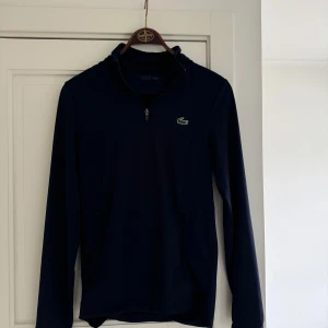 Mörkblå lacoste tröja  - Säljer en stilren mörkblå tröja från Lacoste Sport med dragkedja vid halsen och det klassiska krokodilemblem på bröstet. Tröjan har långa ärmar och är perfekt för en sportig look.