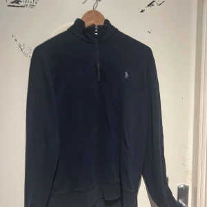 Mörkblå tröja från Ralph Lauren - Säljer en stilren mörkblå tröja från Ralph Lauren med halv dragkedja och det klassiska broderade logotypen på bröstet. Perfekt för en avslappnad men ändå elegant look. Jag säljer den för 800kr. Den är i väldigt fint skick och har inga problem. Säljer då jag har inte haft användning av den.