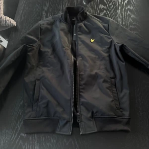 Svart bomberjacka från Lyle & Scott - Snygg svart bomberjacka från Lyle & Scott med dragkedja och ribbade muddar. Jackan har en gul logotyp på bröstet och är perfekt för en stilren look. Den har långa ärmar och en klassisk design.