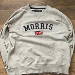 Grå sweatshirt från Morris - Säljer en grå sweatshirt från Morris med texten 'Morris' och en brittisk flagga på framsidan. Tröjan har långa ärmar och en rund halsringning. Perfekt för en avslappnad stil.
