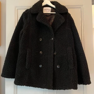 Svart teddyjacka från Stand Studio - Säljer en snygg svart teddyjacka från Stand Studio. Jackan har en dubbelknäppt design med stora knappar och är perfekt för kyligare dagar. Den är långärmad och har en mysig, fluffig textur som ger en trendig look.