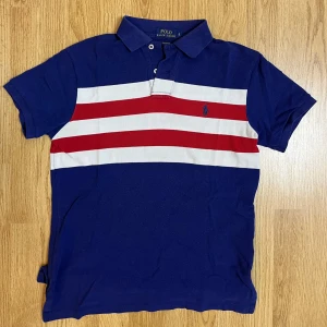   Polo Ralph Lauren pikétröja   - Säljer en snygg blå pikétröja från Ralph Lauren med vita och röda ränder över bröstet. Kom med frågor och bud😁