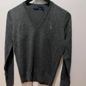 Grå tröja från Ralph Lauren - Säljer en stilren grå tröja från Ralph Lauren med v-ringning och broderad logga på bröstet. Tröjan har långa ärmar och ribbade muddar vid ärmslut och nederkant. Har lagat den lite