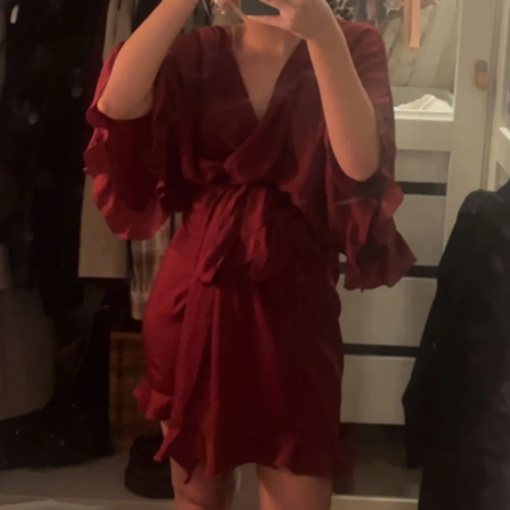 Snygg vinröd satin morgonrock med volangdetaljer och knytband i midjan. Klänningen har en v-ringad hals och trekvartsärmar, perfekt för en elegant look.. Muu.