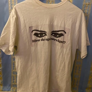 Vit t-shirt med tryck - Vit t-shirt med ett tryck av ögon och texten 'Bliver du også overvæget?' på framsidan. T-shirten har korta ärmar och är tillverkad i bomull. Perfekt för en avslappnad stil.