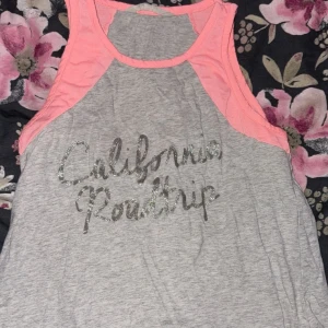 Grå och rosa linnetopp med text - Säljer en grå och rosa linnetopp med texten 'California Roadtrip' i glittriga bokstäver i storlek 170. Toppen har en sportig design med kontrasterande rosa detaljer vid ärmhålen och halsringningen.