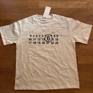 Vit t-shirt Maison Margiela med tryck - Snygg vit t-shirt från Maison Margiela med det ikoniska siffertrycket och en markerad 6:a på bröstet. Klassisk passform, rund hals och korta ärmar. Tillverkad i mjuk bomull som känns skön mot huden. Perfekt för dig som gillar minimalistisk och stilren design.
