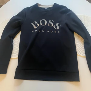 Svart sweatshirt från Hugo Boss - Snygg svart sweatshirt från Hugo Boss med stor BOSS-logga i vitt framtill. Klassisk rund halsringning och långa ärmar. Perfekt för en clean och stilren look, passar till jeans eller joggers.
