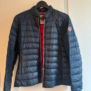 Mörkblå Colmar pufferjacka - Snygg mörkblå pufferjacka från Colmar med röd insida och klassisk logga på ärmen. Jackan har quiltad design, dragkedja framtill och två fickor med dragkedja. Den är deras tunna modell!