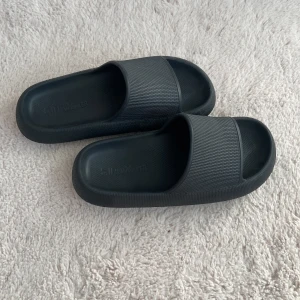 Svarta slides från NewYorker, strl 37 - Svarta slides från NewYorker i storlek 37. Skorna har en chunky, platt sula och bred rem över foten. Tillverkade i syntetmaterial med en clean och minimalistisk look, perfekta för chill dagar eller stranden.