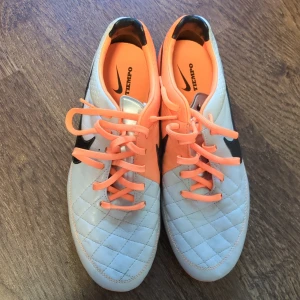 Nike Tiempo fotbollsskor  - Nike Tiempo legend fotbollsskor i ljus grå och orange.  Skorna har snörning, quiltad ovandel och orange sula med dobbar för bra grepp på planen. Jättefin design med klassisk Nike-logga på sidan. 