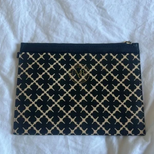 Svart och beige kuvertväska från By Malene Birger - Snygg kuvertväska från By Malene Birger i svart och beige med ikoniskt grafiskt mönster och guldfärgad logga framtill. Väskan har dragkedja upptill och är tillverkad i tåligt syntetmaterial. Perfekt att bära till mobilen, plånboken och småsaker.