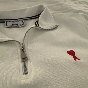 Vit half zip pikétröja från Ami - Vit pikétröja från Ami Alexandre Mattiussi med half zip och klassisk krage med svarta ränder. Tröjan har ett rött broderat hjärta på bröstet och är långärmad. Tillverkad i bomull för en skön och stilren look. Helt ny har aldrig använts