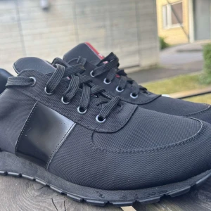 Prada skor  - Snygga svarta sneakers från Prada med klassisk design och diskret röd logga på plösen. Skorna har snörning, rund tå och är tillverkade i slitstark textil med detaljer i skinn. Perfekta för dig som vill ha en stilren och exklusiv look.