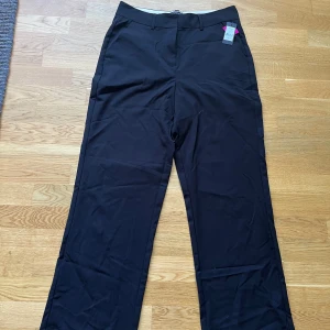 Svarta kostymbyxor från First Class - Klassiska svarta kostymbyxor från First Class Collection med raka ben och bälteshällor. Byxorna har en stilren look, stängs med dragkedja och knapp framtill och har fickor i sidorna.