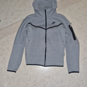 Grå Nike hoodiejacka med dragkedja M - Snygg grå hoodiejacka från Nike med hel dragkedja och svarta detaljer. Jackan har huva, lång ärm och en cool ficka med dragkedja på ärmen. Perfekt för dig som gillar sportig och clean stil. Nike-logga på bröstet och kontrastband längs dragkedjan.