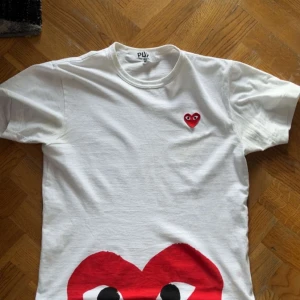 Vit Comme des Garçons Play t-shirt - Säljer en vit t-shirt från Comme des Garçons Play med det ikoniska röda hjärtat med ögon på bröstet och ett stort hjärta nertill. Klassisk passform, rund hals och korta ärmar. Tillverkad i 100% bomull – perfekt för en clean och cool look.