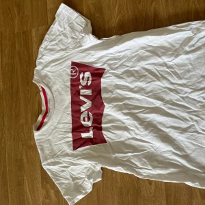 Vit Levi's t-shirt med röd logga - Vit t-shirt från Levi's med klassisk röd logga framtill. T-shirten har rund hals med röd detalj på insidan och är kortärmad. Tillverkad i mjuk bomull, perfekt för en avslappnad och stilren look.