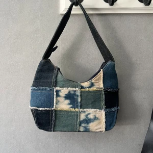 Patchwork axelväska i jeans - Unik axelväska i patchwork-stil med olika nyanser av blått och beige denim. Väskan har fransiga kanter mellan lapparna och en bred axelrem. Stängs med dragkedja upptill och har en cool, retro vibe som sticker ut. Jätte unik och cool! 