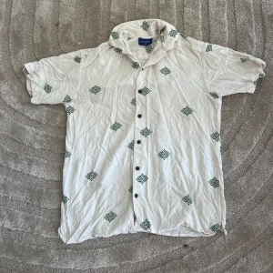 Vit mönstrad skjorta Jack & Jones - Vit skjorta från Jack & Jones med grönt grafiskt mönster över hela plagget. Skjortan har korta ärmar, klassisk krage och svarta knappar framtill. Perfekt för en chill sommarlook.