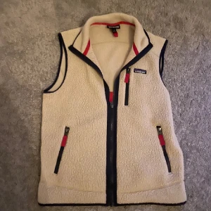 Beige fleeceväst från Patagonia - Säljer en beige fleeceväst från Patagonia med svarta detaljer och röda dragkedjor. Västen har hög krage, tre fickor med dragkedja och klassisk Patagonia-logga på bröstet. Perfekt för lager-på-lager och chill vardagsstil.