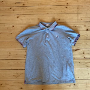 Grå Moncler pikétröja med logga - Snygg grå pikétröja från Moncler med klassisk krage och knäppning framtill. Tricolore-detaljer i rött, vitt och blått på krage och ärmslut. Broderad Moncler-logga på bröstet. Perfekt för en clean och sportig look.