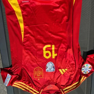 Spansk landslagströja Adidas röd - Säljer en röd fotbollströja från Adidas, officiella spanska landslaget med nummer 19 i gult. Tröjan har gula detaljer längs sidorna och axlarna, samt flera broderade märken och patchar på bröstet och ärmarna. Tillverkad i lätt och ventilerande material. Replika, endast handtvätt gäller