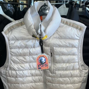 Ljusbeige dunväst från Parajumpers - Snygg ljusbeige dunväst från Parajumpers med quiltad design och hög krage. Västen har dragkedja framtill, tre fickor med dragkedja och en ikonisk gul detalj vid halsen. Perfekt att slänga över en hoodie för en cool look.