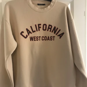 Beige sweatshirt California Boohoo - Beige sweatshirt från Boohoo med trycket 'California West Coast' i svart och rött framtill. Tröjan har rund halsringning och långa ärmar, perfekt för en avslappnad och trendig look.