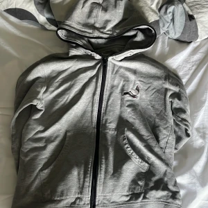 Grå hoodiejacka från Staple med duva - Grå hoodiejacka från Staple med broderad duva på bröstet och logga på ärmen. Jackan har dragkedja framtill, huva och två stora fickor. Tillverkad i mjuk bomull, perfekt för chill dagar eller streetwear-vibe.