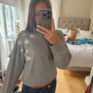 Grå hoodie med stjärnor  - Grå hoodie från Gina Tricot med vita stjärnor på axeln och en stor stjärna på ryggen. Tröjan har huva, långa ärmar och är i mjukt bomullsmaterial. Perfekt för chill dagar och att ha på kvällskvisten när man sitter ute!